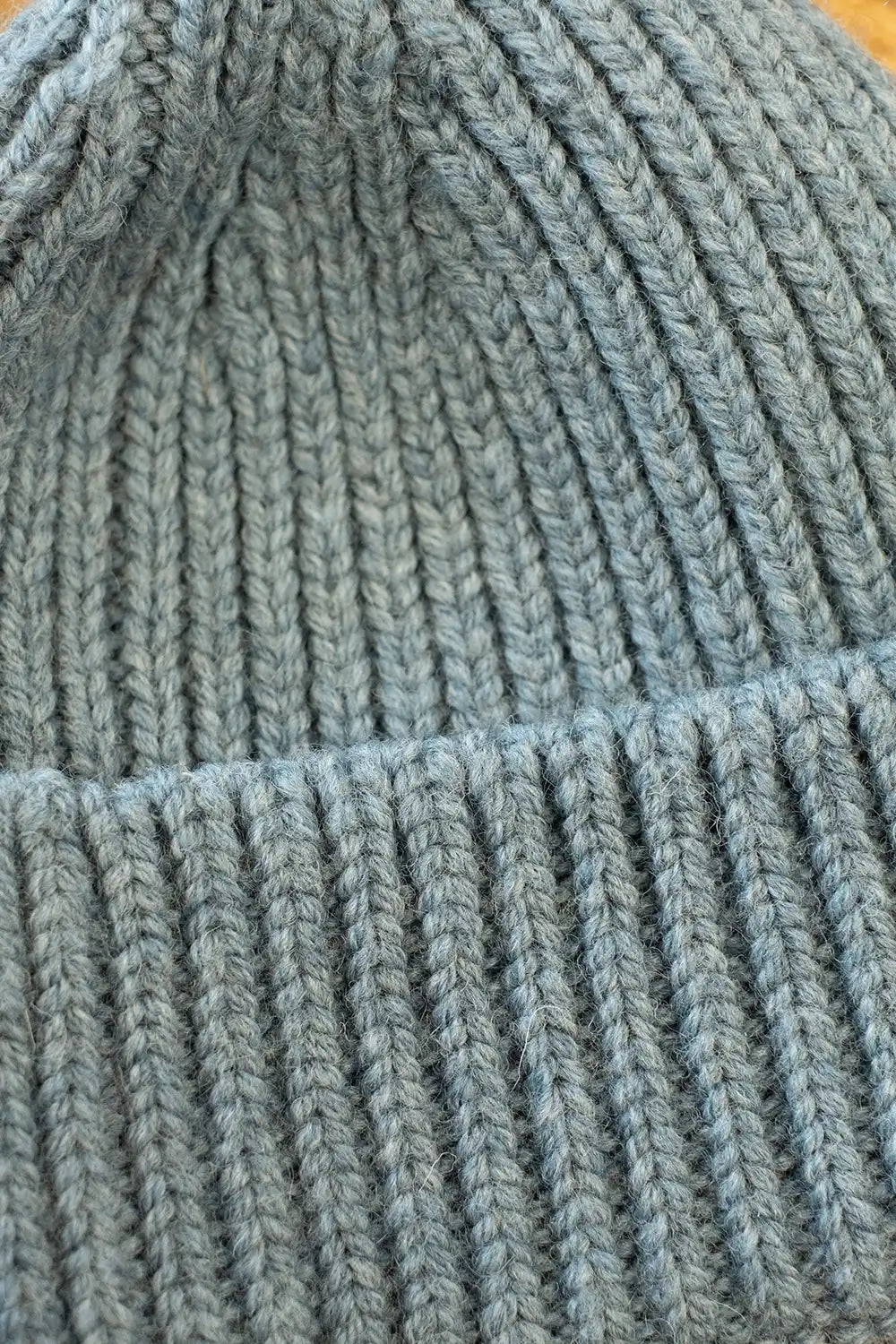 Close-up of a chunky knit beanie hat in a dusty slate blue color, showing the detailed ribbed texture and folded brim.