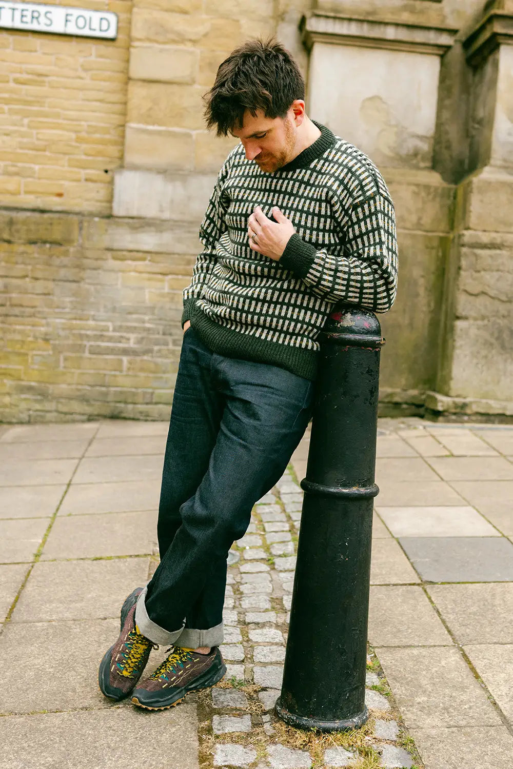 A man wearing a patterned knit jumper, dark blue jeans, and hiking boots leans against a black metal bollard on a cobblestone street in front of a brick building.