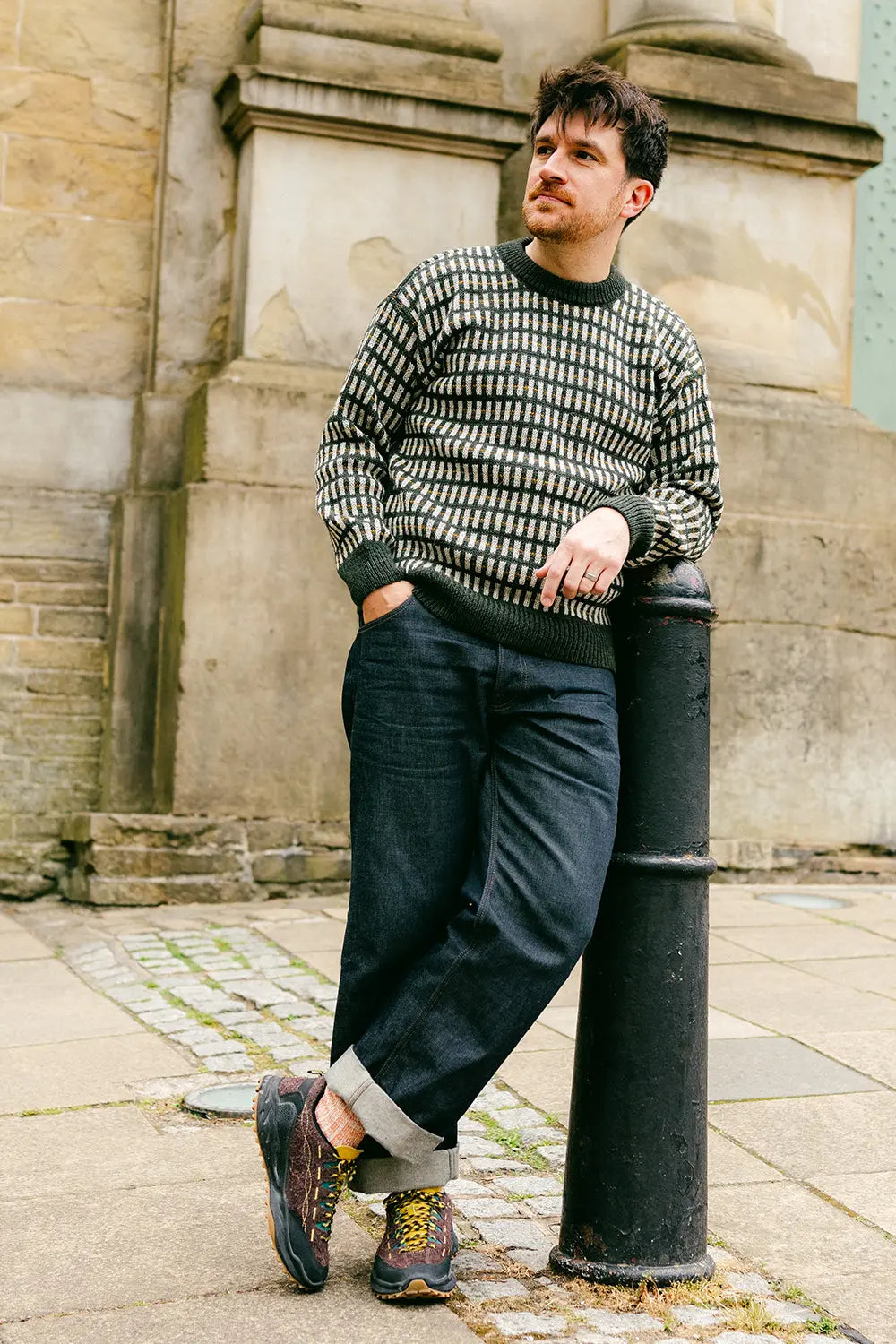 A man wearing a patterned knit jumper, dark blue jeans, and brown hiking boots leans against a black metal bollard in front of a stone building.