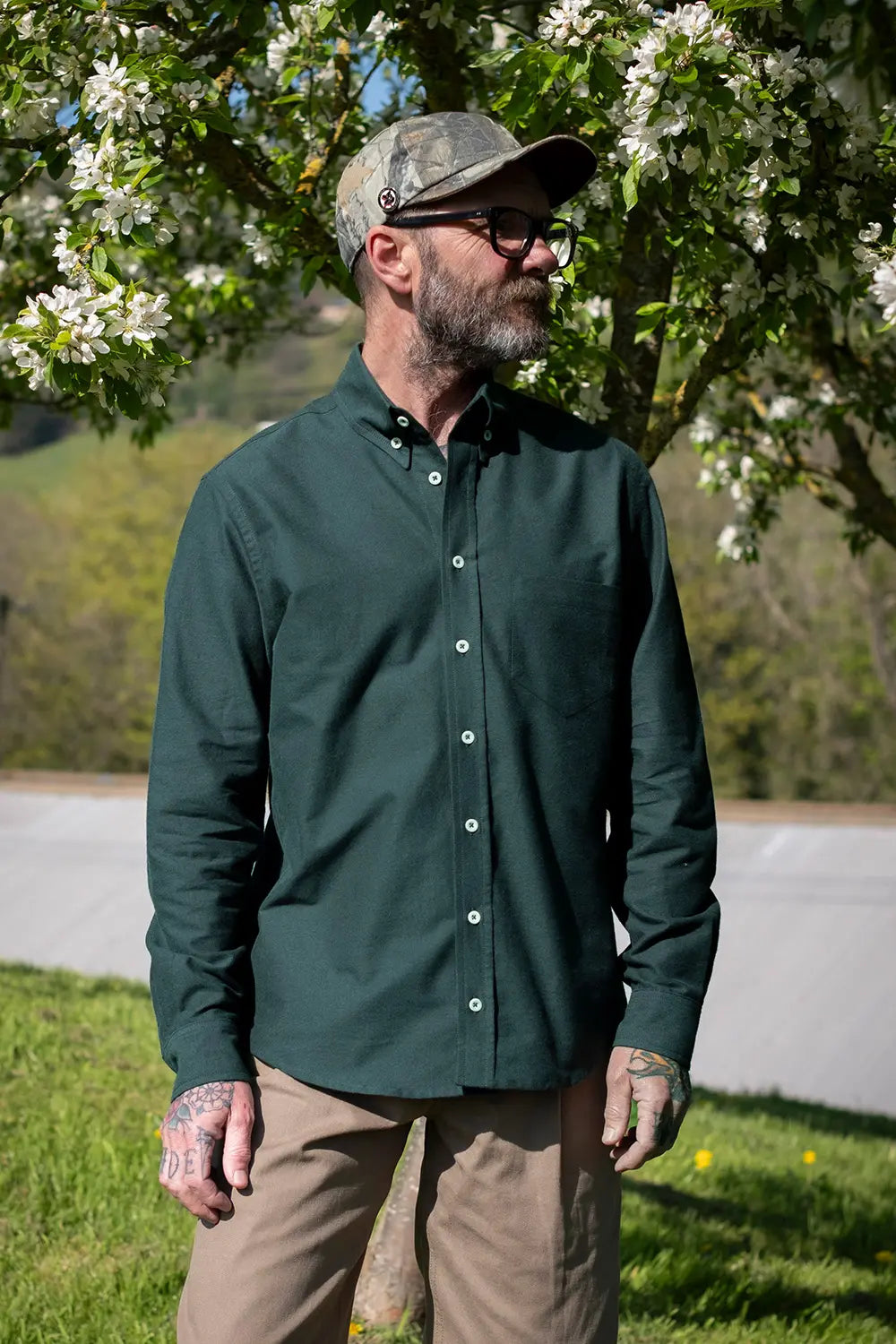 A man wearing a dark green long-sleeve button-down oxford shirt, tan pants, and a camouflage flat cap stands outdoors under a flowering tree.