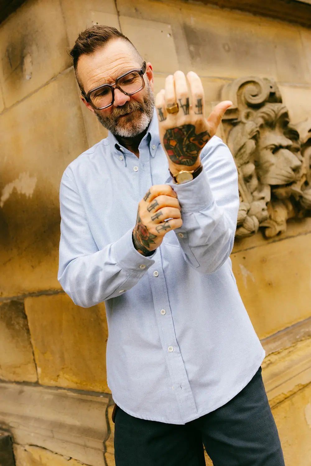 A man with glasses, a beard, and tattoos poses in front of a stone wall, holding up his hand to show a large, custom black ink tattoo on the back of his hand. He is wearing a light blue button-down shirt and dark trousers.