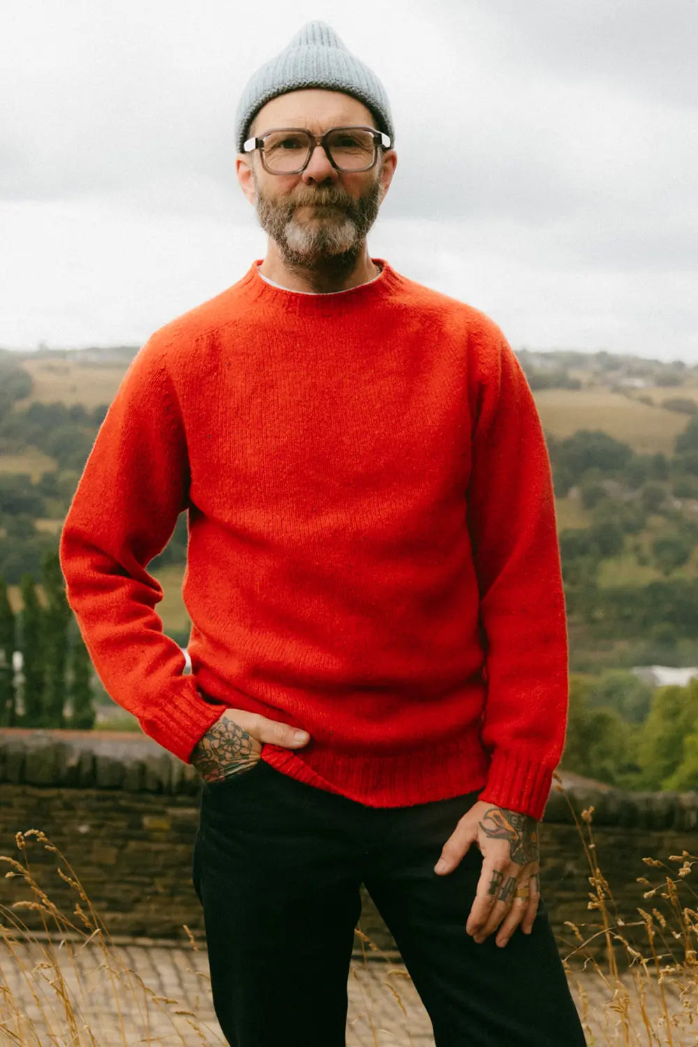 A man wearing a bright red knit crewneck jumper, a light gray beanie, and glasses, standing outdoors against a blurred landscape background.