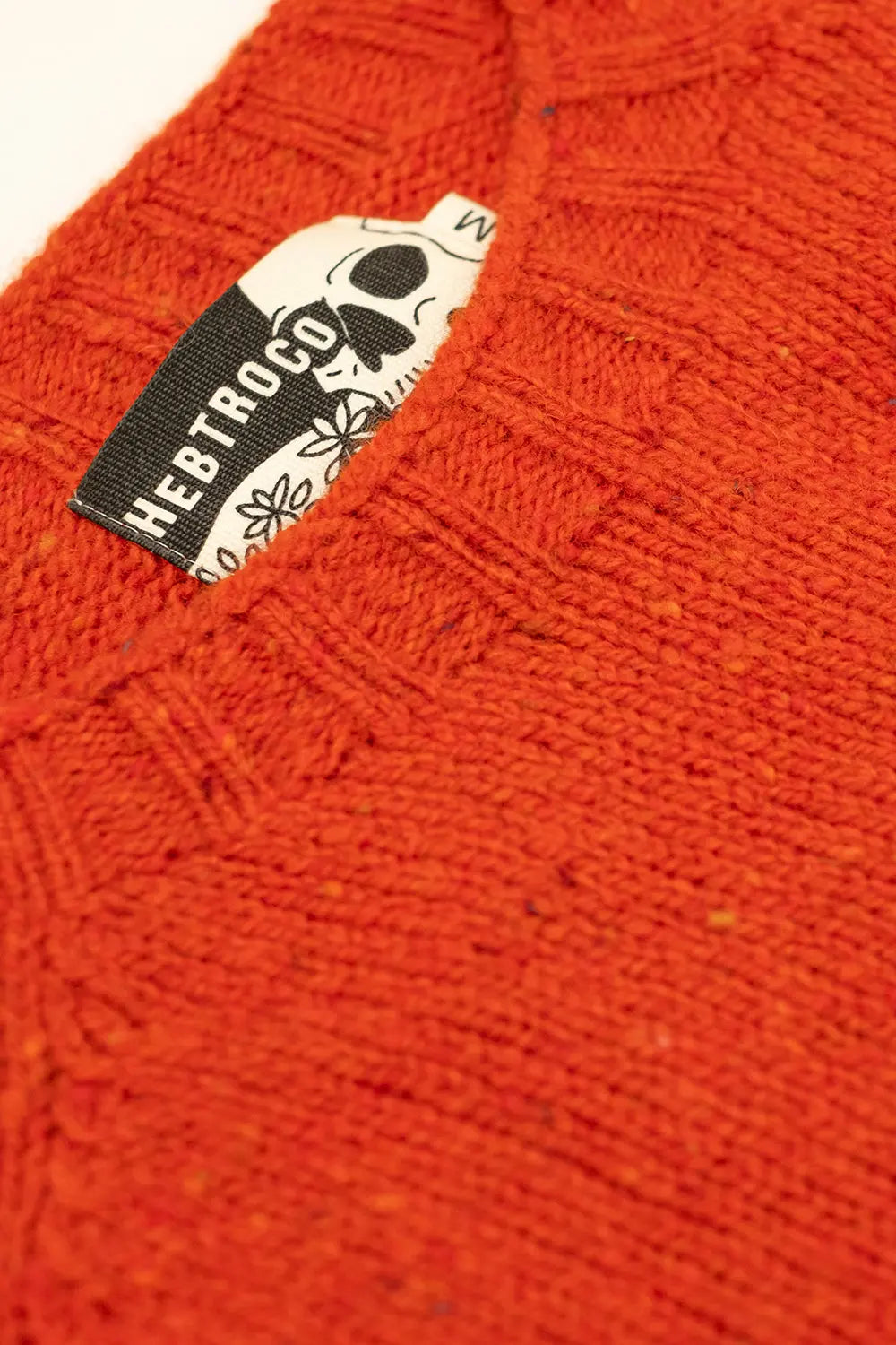 Close-up of a textured knit sweater in a burnt orange color, featuring a black and white brand label that reads 'HEBTROCO' with a skull graphic.
