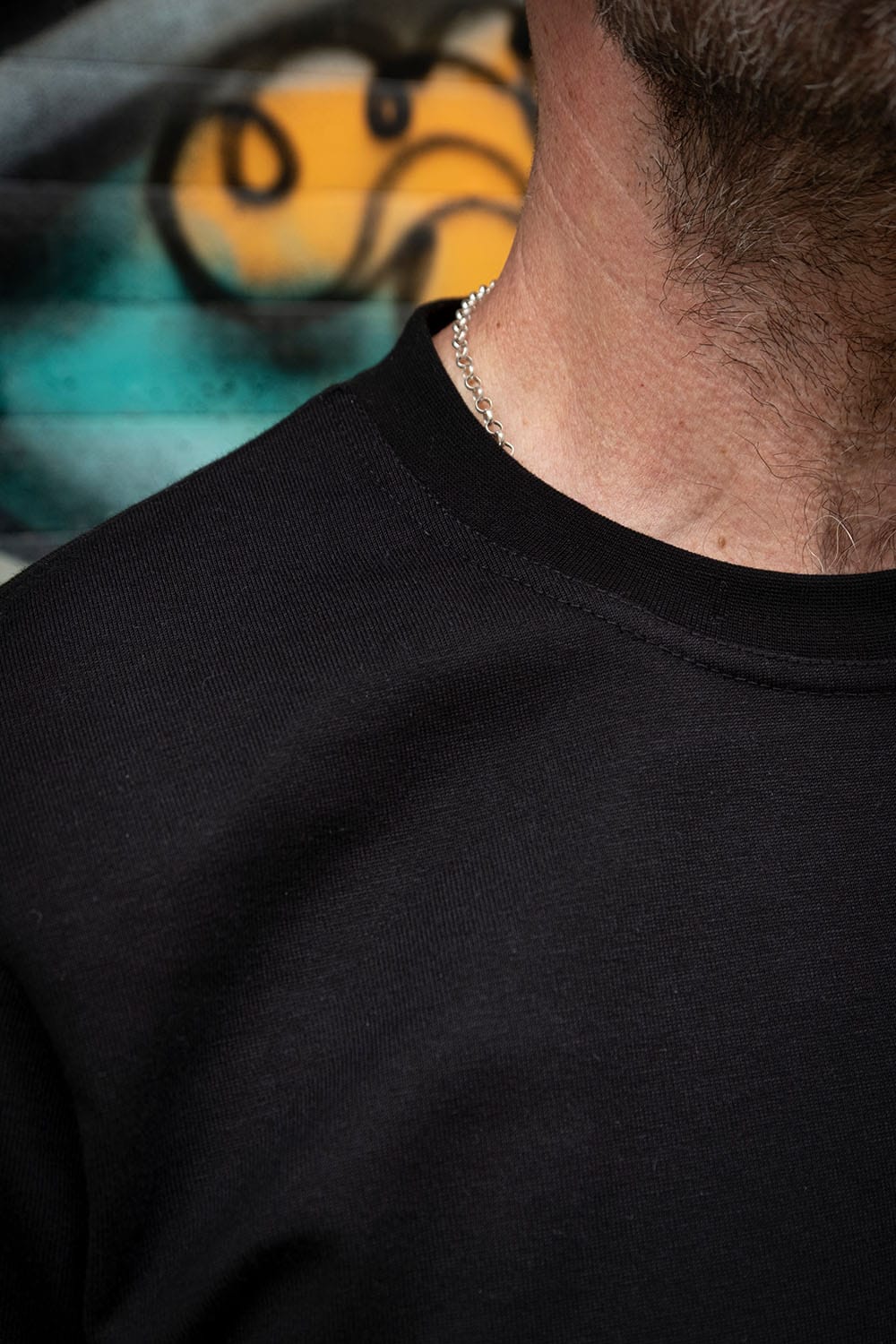 Close-up of a person wearing a black crewneck sweatshirt, showing the ribbed fabric texture and collar, with a blurred graffiti background.