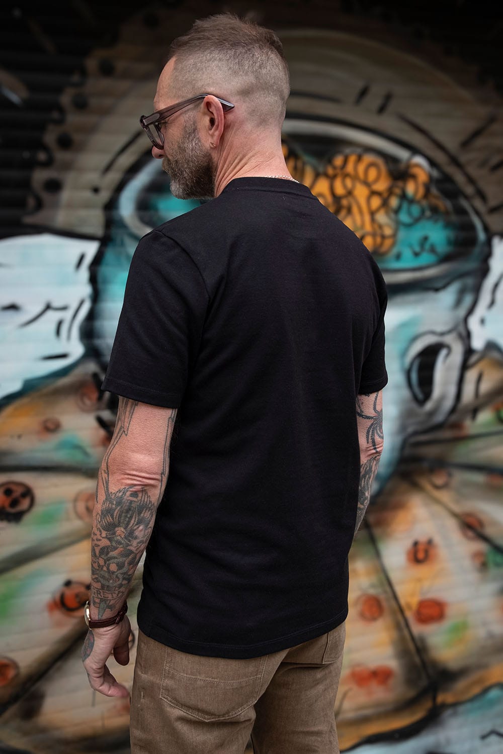 A man with tattoos and glasses, seen from behind, wearing a black short-sleeve t-shirt and brown pants, standing in front of a colorful graffiti mural.