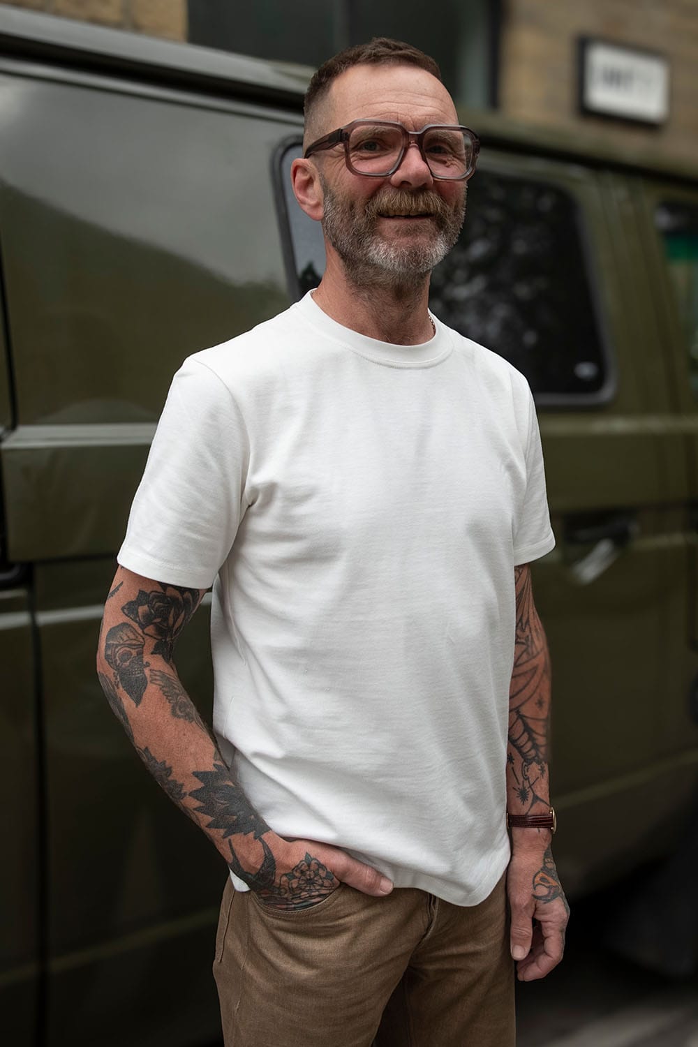 A man with glasses, a beard, and tattoos wearing a plain white crewneck t-shirt and brown trousers, standing in front of an olive green van.