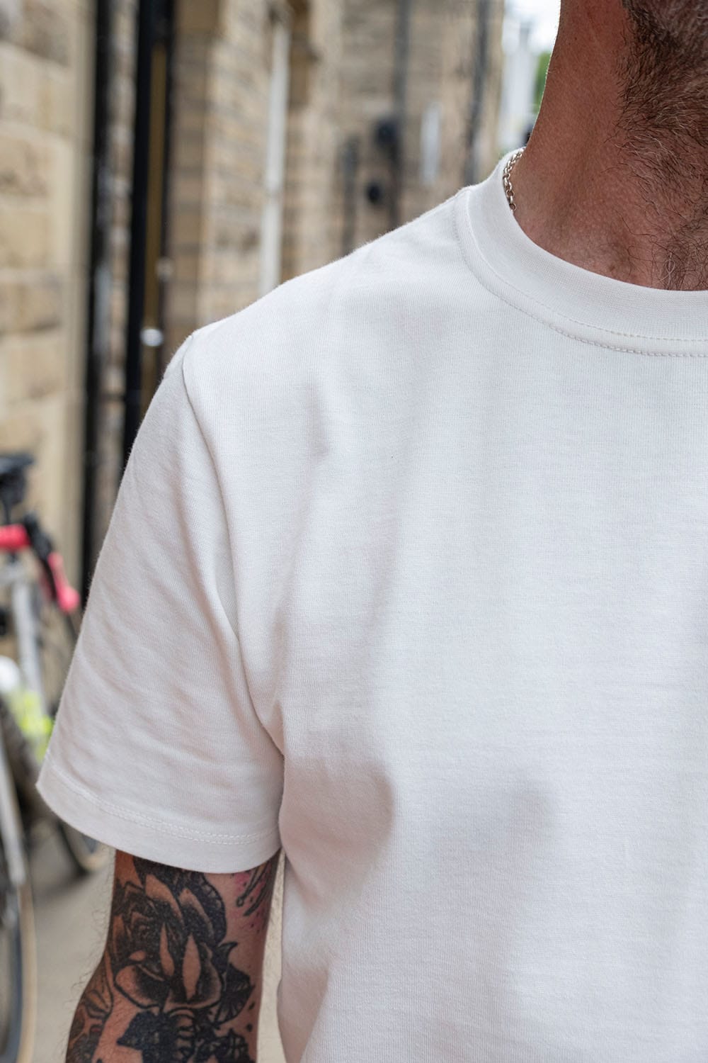A close-up shot of a man wearing a plain white short-sleeve crewneck t-shirt, with a tattooed arm visible on the left side and a blurred outdoor street background.