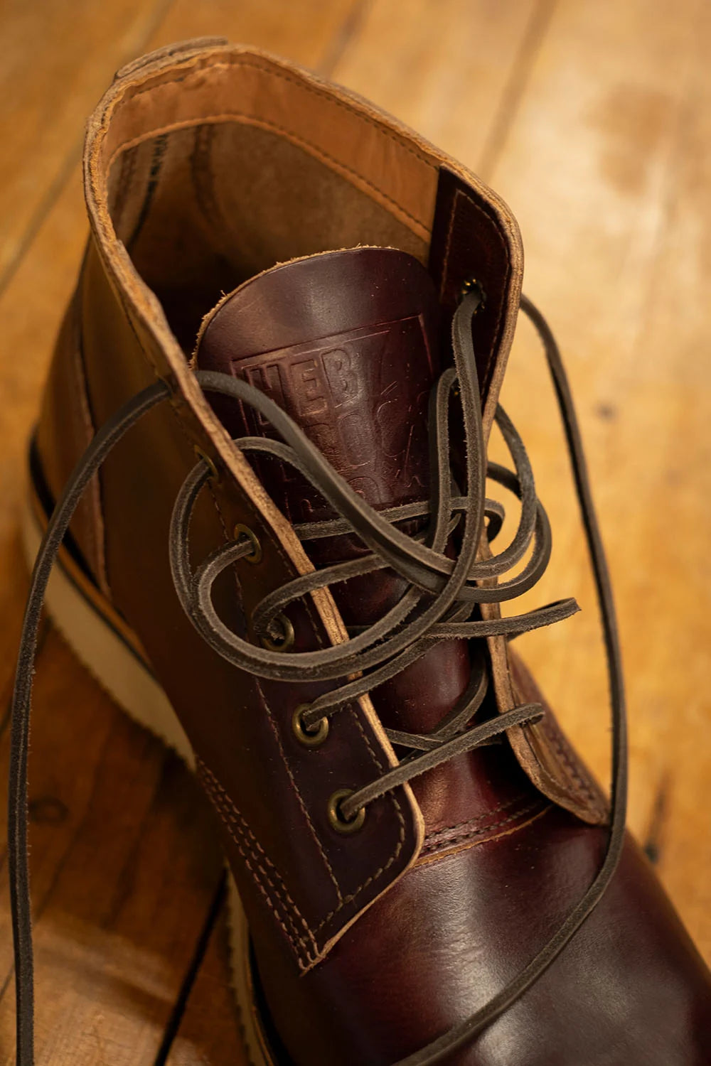 A close-up of a dark brown leather lace-up boot with dark laces, resting on a warm wooden floor. The embossed brand name 'HEBTROCO' is visible on the tongue.