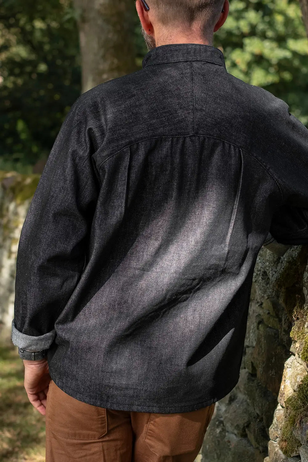 A man viewed from behind wearing a dark charcoal gray chambray button-down shirt and brown pants, standing outdoors next to a stone wall.
