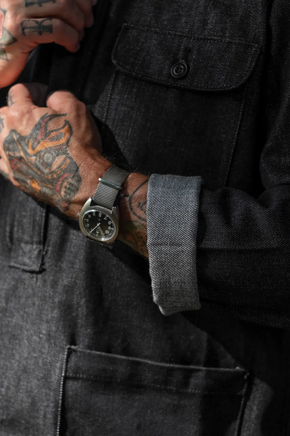 A close-up of a person wearing a dark charcoal gray canvas utility jacket, showing a tattooed hand adjusting the collar and a silver-toned dial watch with a gray mesh strap on the wrist.