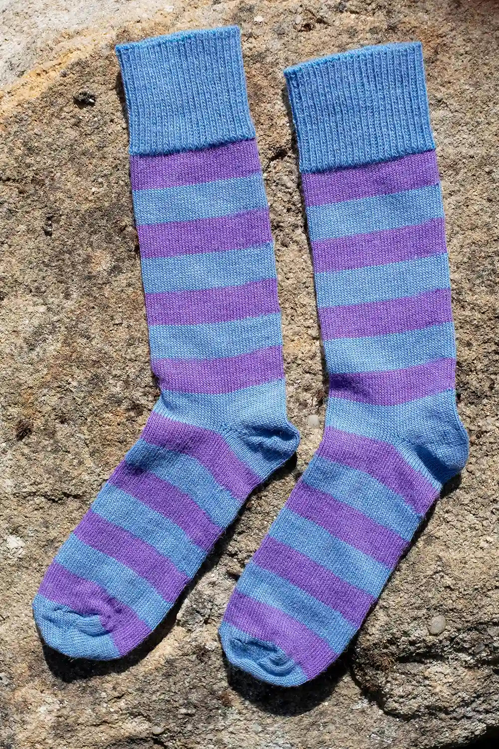 A pair of knee-high striped socks in shades of blue and purple, laid flat on a textured stone surface.