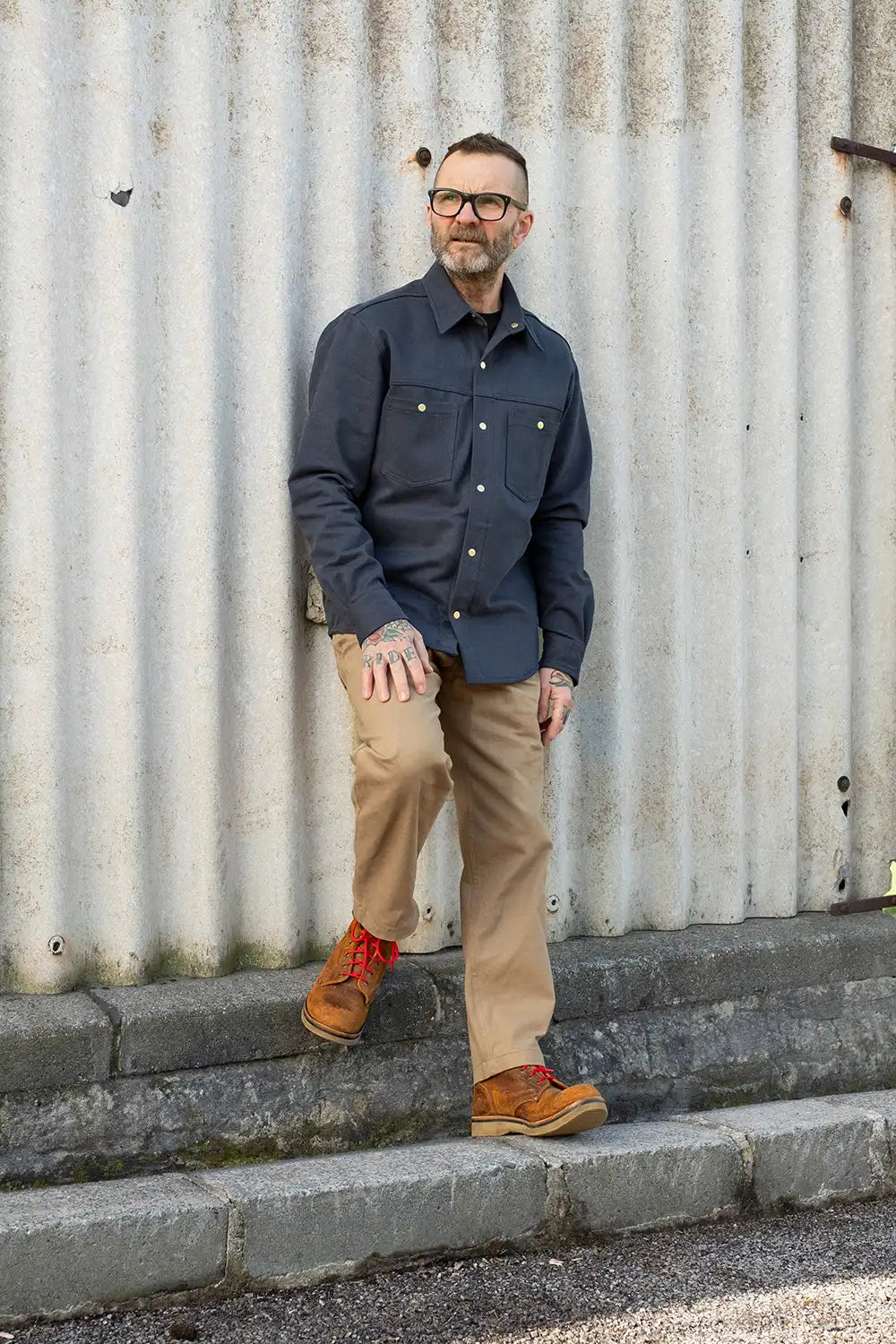 A man wearing a dark bedford cord ironshirt, tan pants, and brown boots stands on stone steps against a corrugated metal wall.