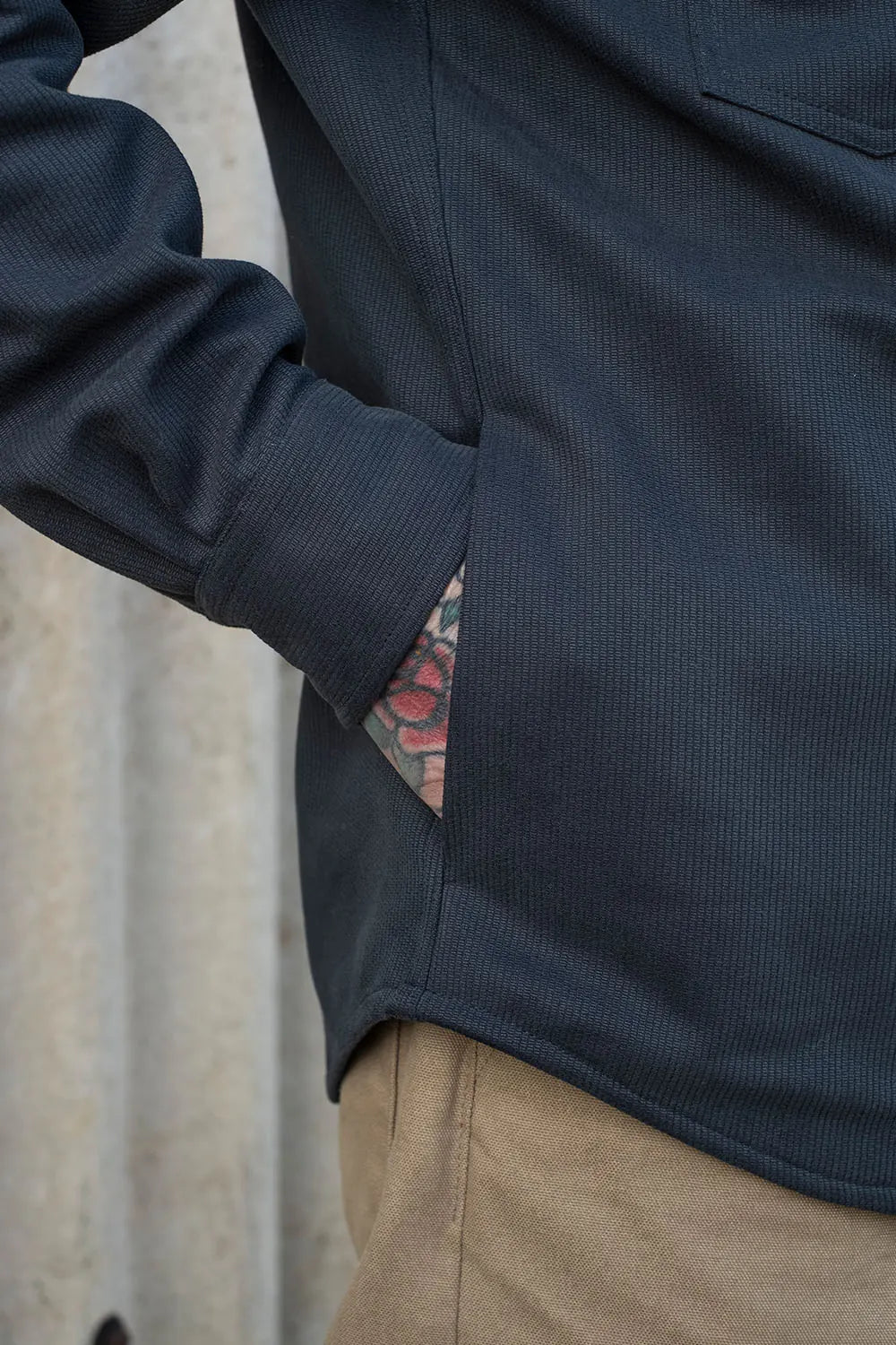 Close-up of a person wearing a dark navy bedford cord iron shirt with a side pocket, showing a tattooed arm in the pocket and tan pants against a light textured wall.