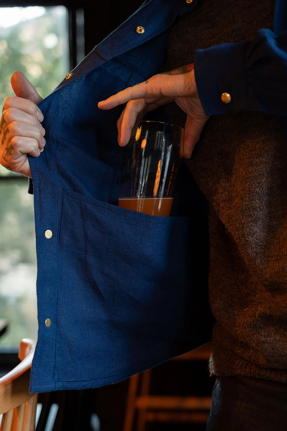 A close-up of a person wearing a dark blue button-up shirt and brown sweater, holding open the shirt to show a clear glass tumbler tucked into the front pocket.