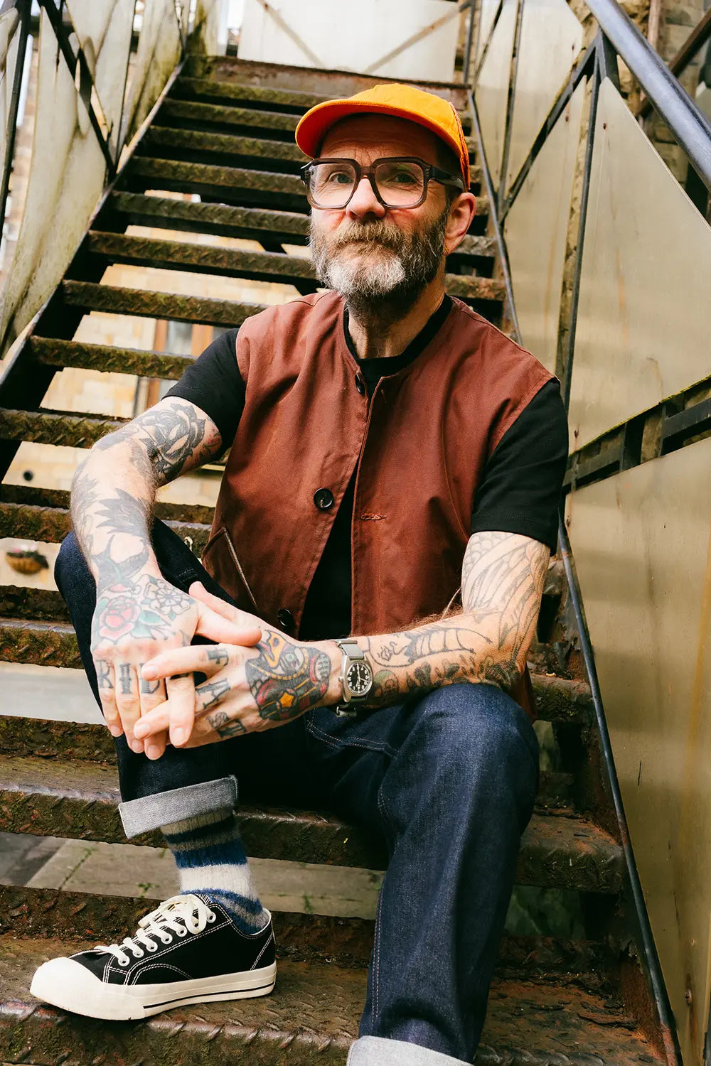 A man with a beard, glasses, and tattoos sits on an outdoor metal staircase. He is wearing a rust-brown vest over a black t-shirt, dark blue jeans, black canvas sneakers, and an orange baseball cap.