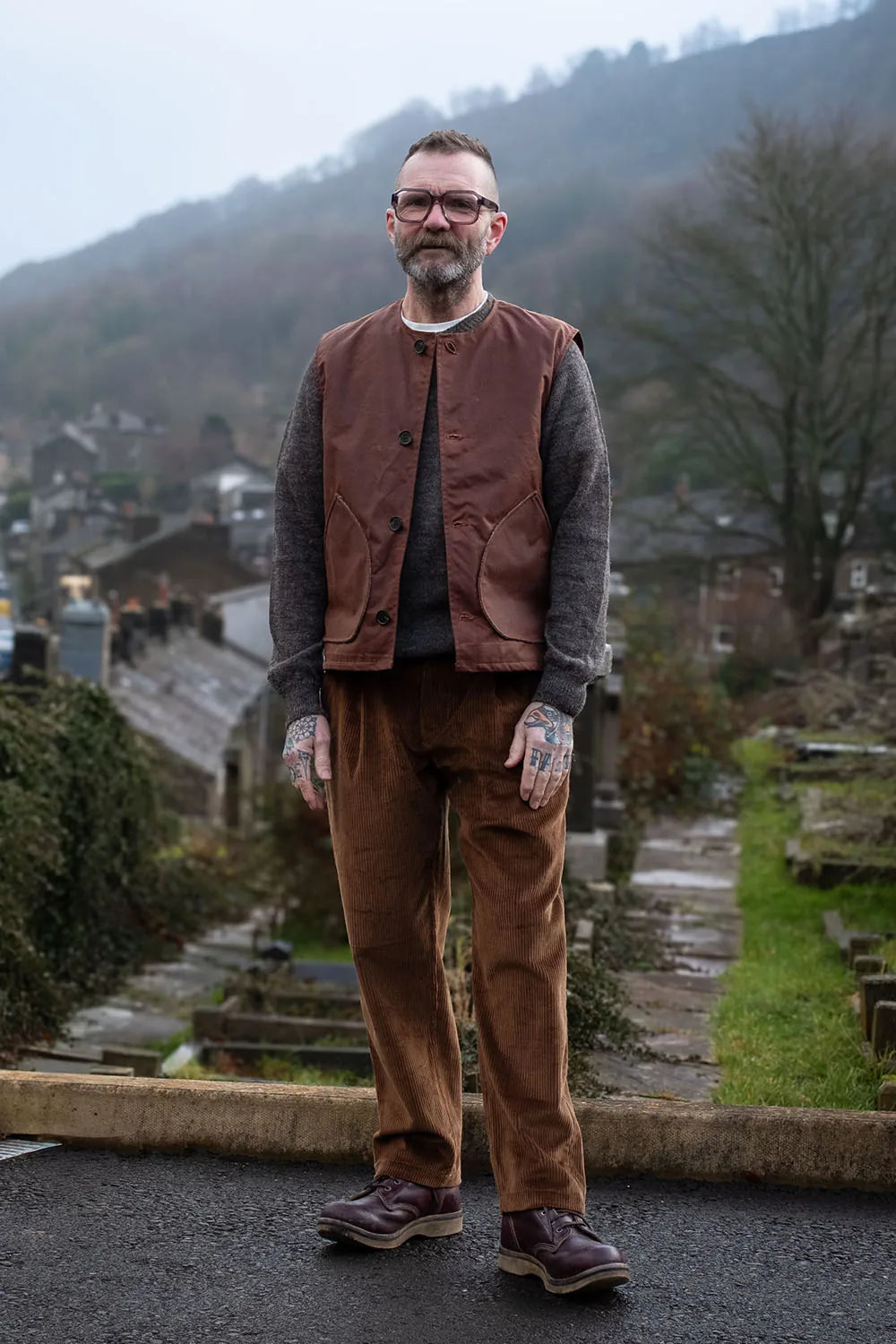 Ed wearing a Rascal vest over a dark gray jumper, brown corduroy trousers, and brown leather shoes, standing outdoors in a rural setting with a hillside village and trees in the background.