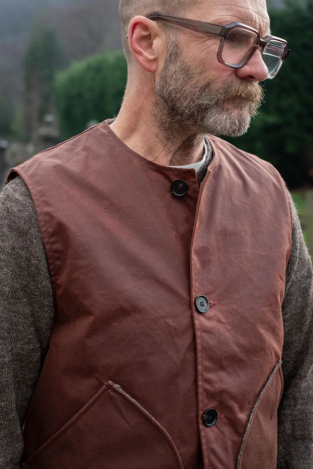 A close-up of a man wearing a brown waxed cotton utility vest over a textured gray sweater, with a blurred outdoor background.