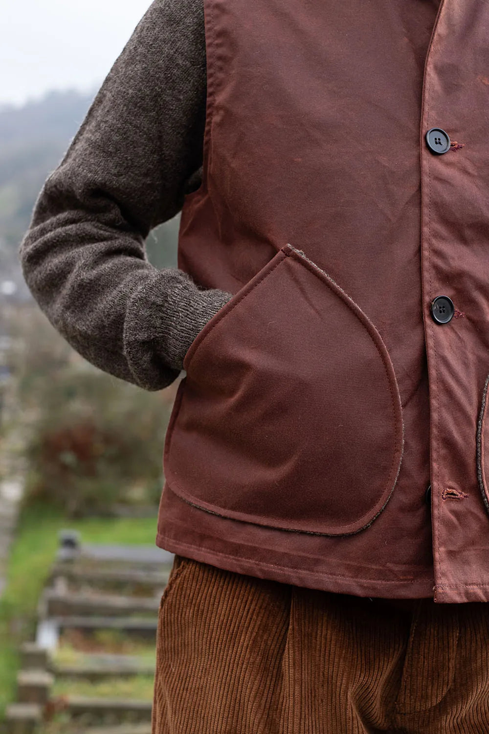 A close-up of a person Rascal waxed cotton vest over a dark brown knit sweater and brown corduroy trousers, with an outdoor blurred background.
