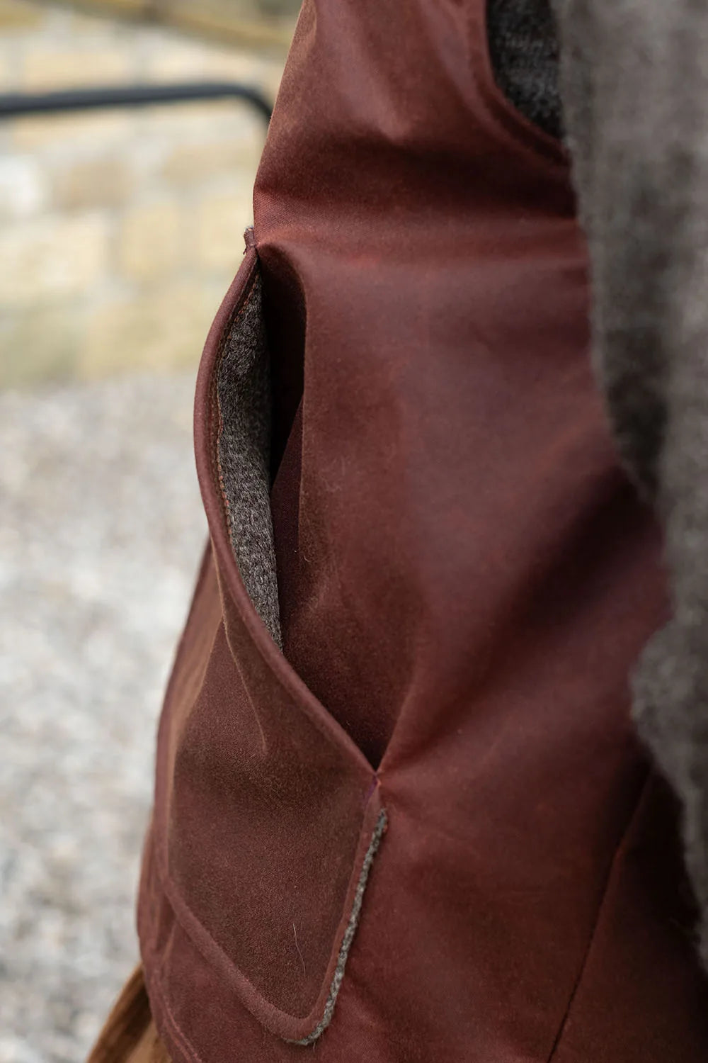 Close-up of a rascal vest with a side pocket showing a gray textured lining, set against a blurred outdoor background.