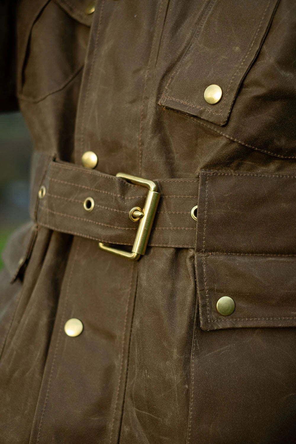Close-up of a dark brown waxed canvas jacket with a matching belt and brass buckle, showing the texture and details of the fabric and metal hardware.