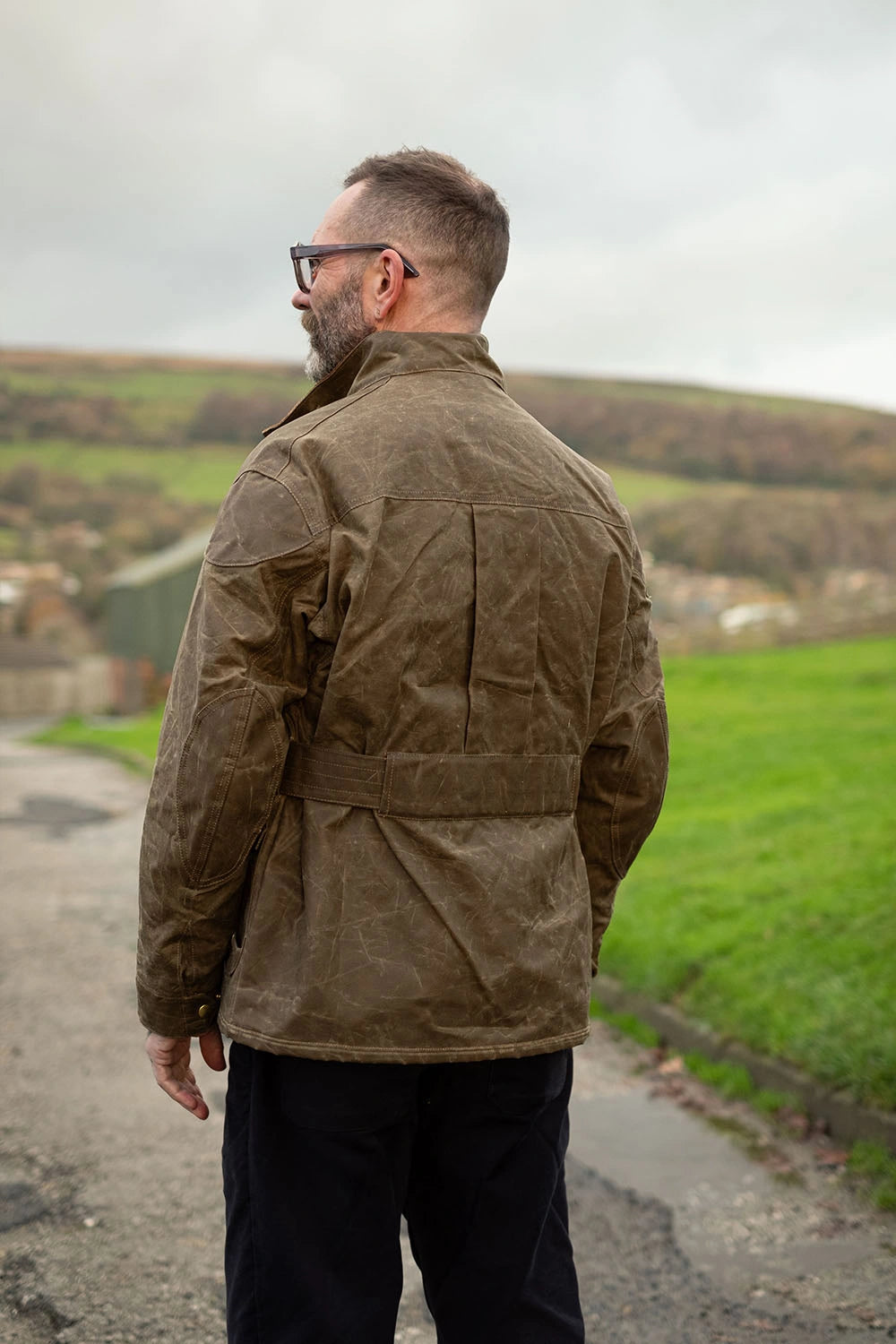 A man wearing a brown waxed canvas roadrunner jacket and black trousers, photographed from behind in an outdoor rural setting with green fields and hills under an overcast sky.