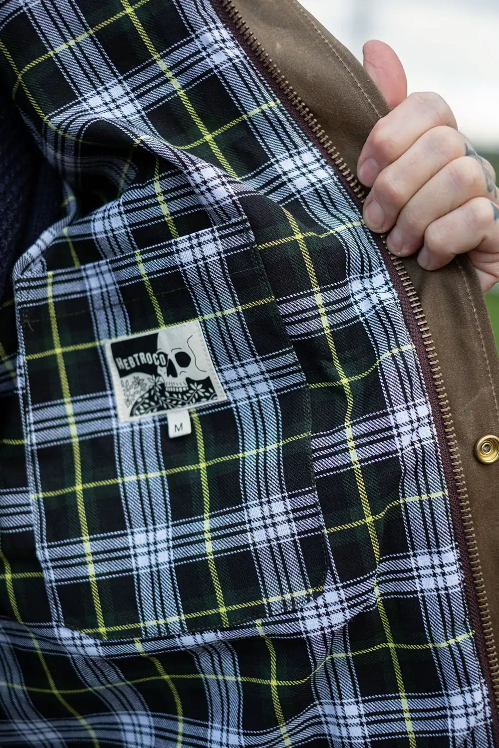 Close-up of a person's hand holding open a brown jacket to show the interior lining, which features a black, white, and lime green plaid pattern and a visible 'Wealth' brand patch.