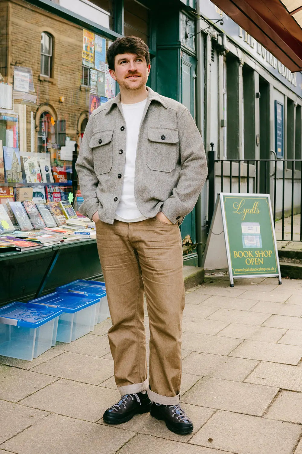 A man wearing a USB37 wool overshirt over a white t-shirt, tan trousers, and black sneakers, standing on a paved street in front of a bookshop.