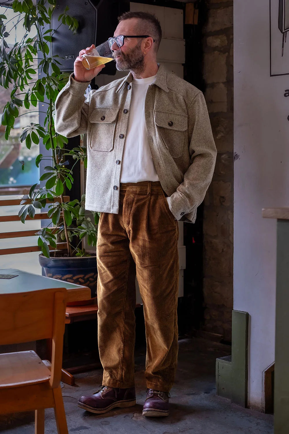 A man wearing a USB37 wool overshirt over a white t-shirt, brown corduroy trousers, and brown leather shoes, standing in a rustic indoor setting and drinking from a glass.
